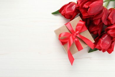 Beautiful gift box with bow and red tulip flowers on white wooden background, flat lay. Space for text