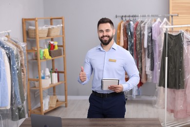 Photo of Dry-cleaning service. Happy worker with notebook showing thumb up in workplace indoors