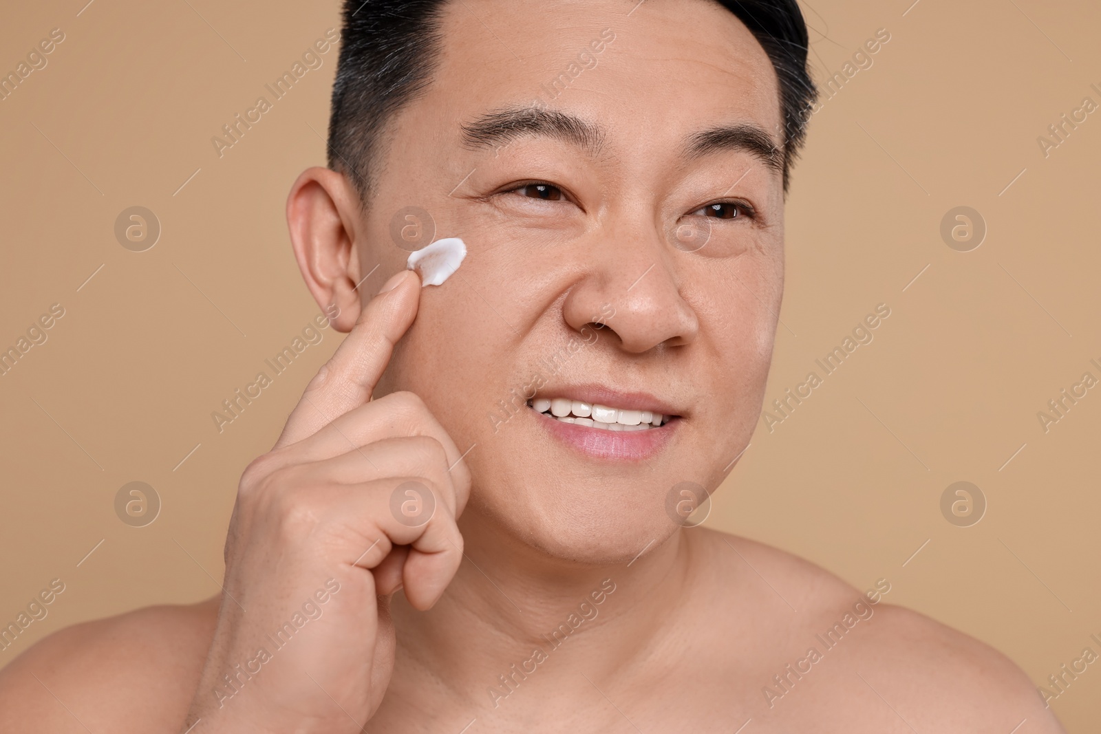 Photo of Handsome man applying cream onto his face on light brown background, closeup