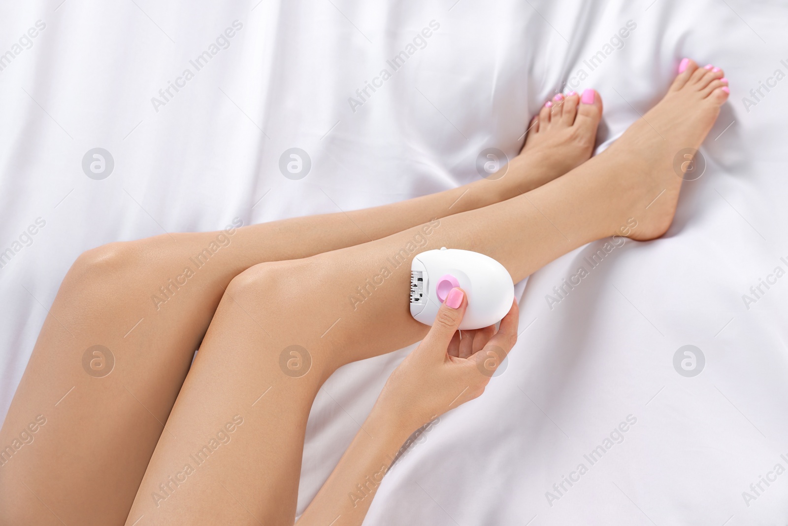 Photo of Young woman epilating her legs on bed, above view