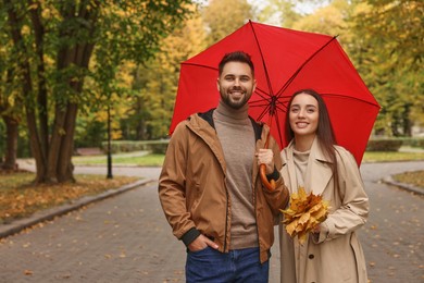 Happy young couple with red umbrella spending time together in autumn park, space for text