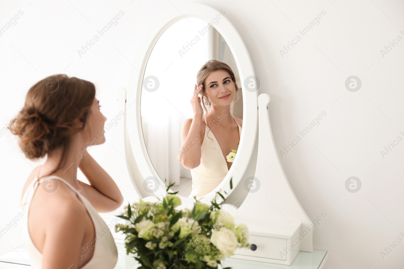 Photo of Young bride in wedding dress with beautiful bouquet near mirror indoors