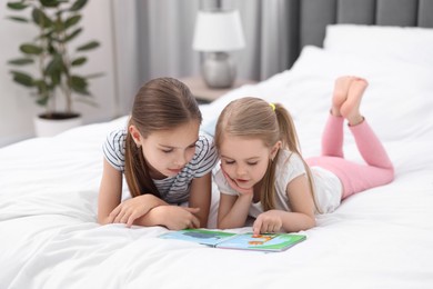 Photo of Cute little sisters reading book together on bed at home