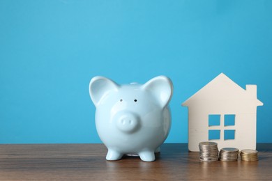 Photo of House model, piggy bank and stacked coins on wooden table against light blue background, space for text