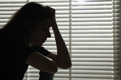 Photo of Silhouette of sad young woman near closed blinds indoors, space for text