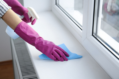 Photo of Woman cleaning window sill with rag and detergent indoors, closeup