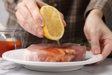 Photo of Woman squeezing lemon onto raw marinated meat at white marble table, closeup