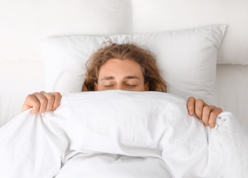 Photo of Young man covering his face with blanket while sleeping on pillow, top view. Bedtime