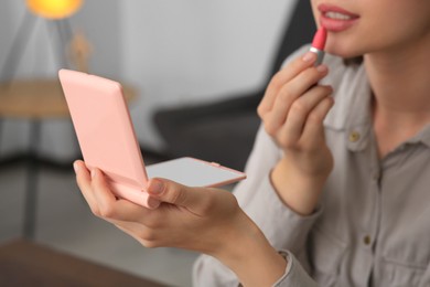 Photo of Young woman with cosmetic pocket mirror applying lipstick indoors, closeup