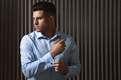 Photo of Handsome stylish man putting on cufflink near wooden wall. Space for text