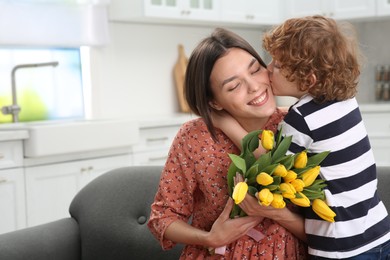 Photo of Little son kissing and congratulating his mom with Mother`s day at home, space for text. Woman holding bouquet of yellow tulips
