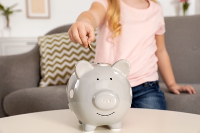 Photo of Cute girl putting coin into piggy bank at table, closeup. Saving money