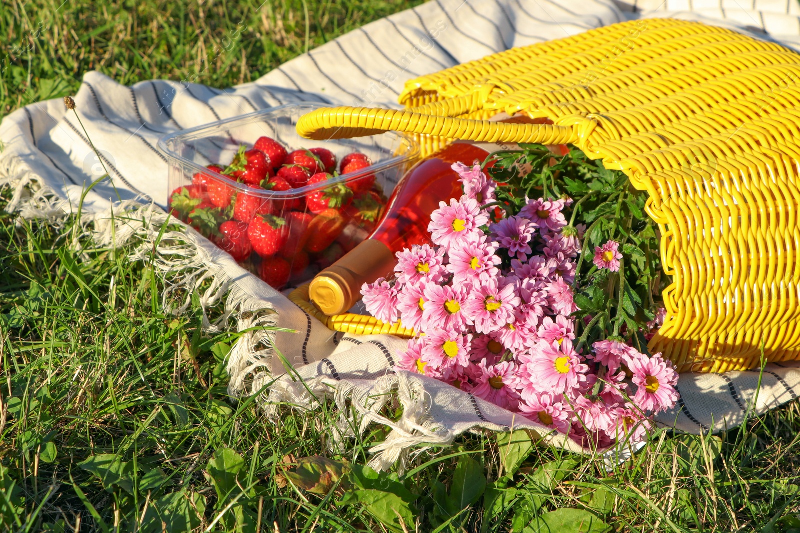 Photo of Yellow wicker bag with beautiful flowers, bottle of wine and strawberries on picnic blanket outdoors