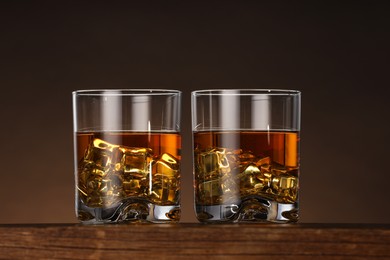Photo of Whiskey with ice cubes in glasses on wooden table against brown background, closeup