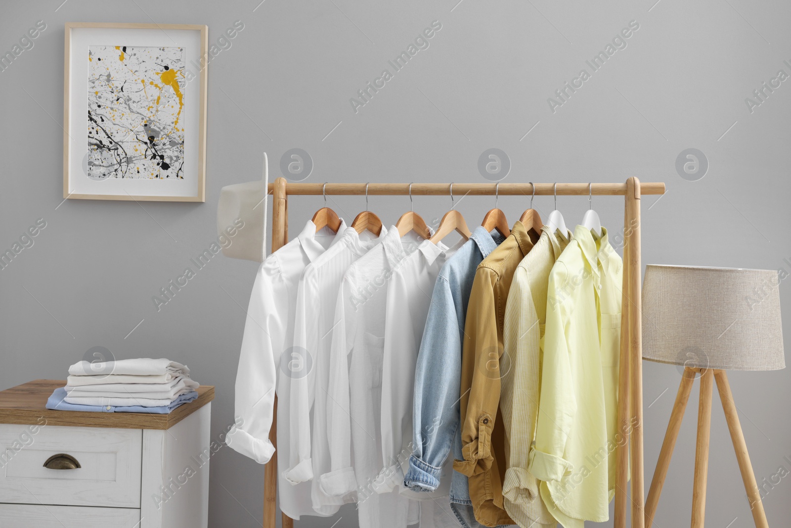 Photo of Rack with different stylish shirts, chest of drawers and lamp near grey wall indoors. Organizing clothes