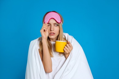 Photo of Young woman in sleeping mask wrapped with blanket holding cup on blue background