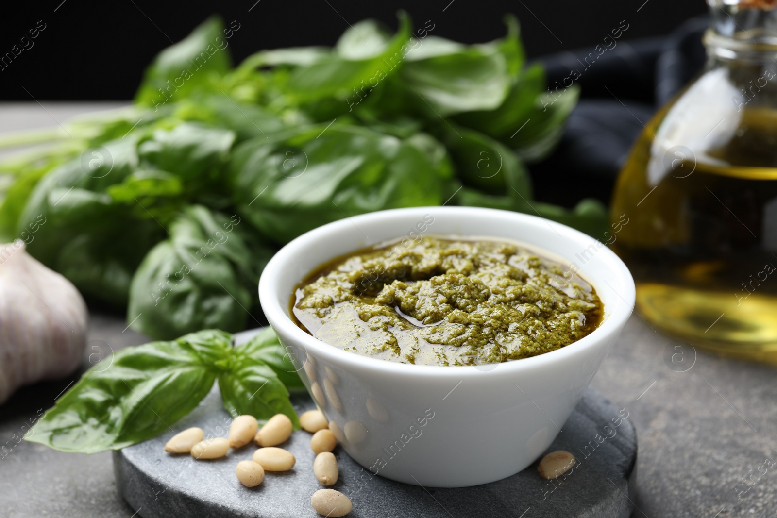 Photo of Tasty pesto sauce in bowl, basil, pine nuts and oil on grey table