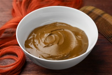 Photo of Bowl of henna cream and red strand on wooden table, closeup. Natural hair coloring
