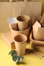 Photo of Eco friendly food packaging. Paper containers, tableware, bag and eucalyptus leaves on pale yellow background