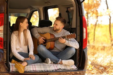 Photo of Young couple with guitar sitting in open car trunk outdoors