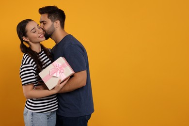 Photo of Man kissing his smiling girlfriend on orange background, space for text. Celebrating holiday