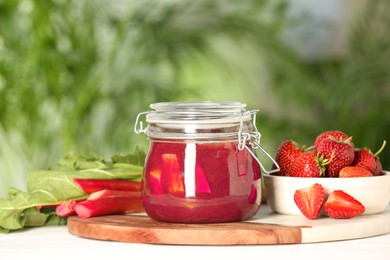 Photo of Jar of tasty rhubarb jam, fresh stems and strawberries on white table against blurred background. Space for text