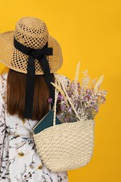 Photo of Woman holding beach bag with beautiful bouquet of wildflowers and book on yellow background, back view