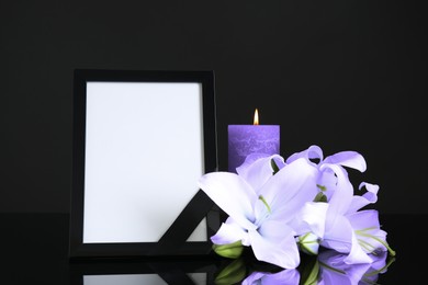 Image of Funeral photo frame with ribbon, violet lily flowers and candle on dark table against black background