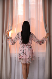 Photo of Young woman wearing floral print dress near window at home, view from back
