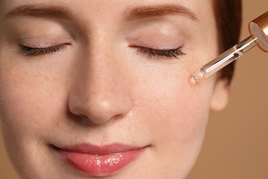 Beautiful woman with freckles applying cosmetic serum onto her face on beige background, closeup