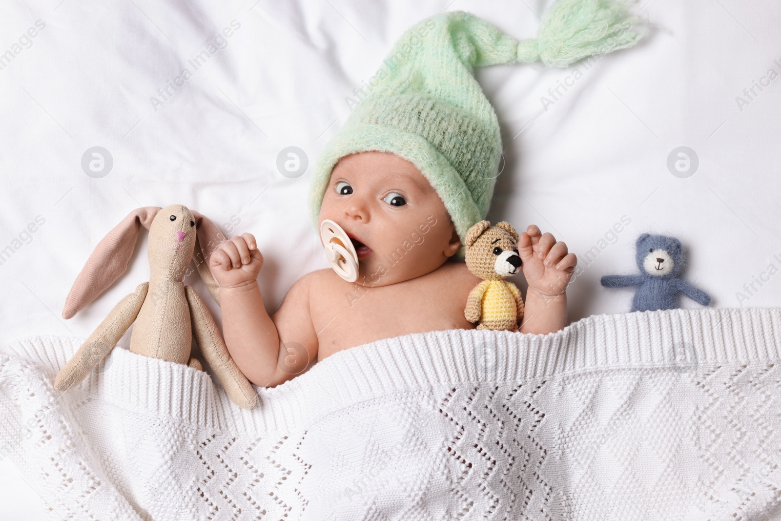 Photo of Cute little baby with toys lying under knitted plaid in bed, top view