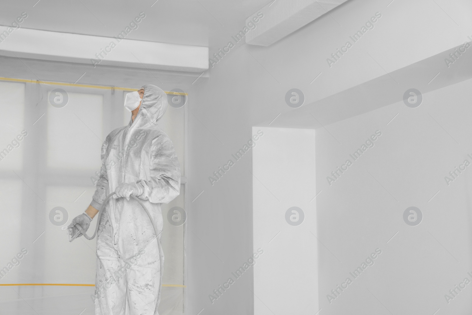 Photo of Decorator in protective overalls painting ceiling with spray gun indoors