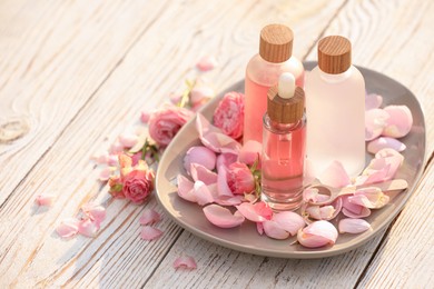 Bottles of rose essential oil and flowers on white wooden table, space for text