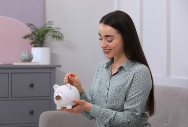 Young woman putting coin into piggy bank at home