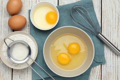 Photo of Whisk, eggs in bowl and flour on white wooden table, flat lay