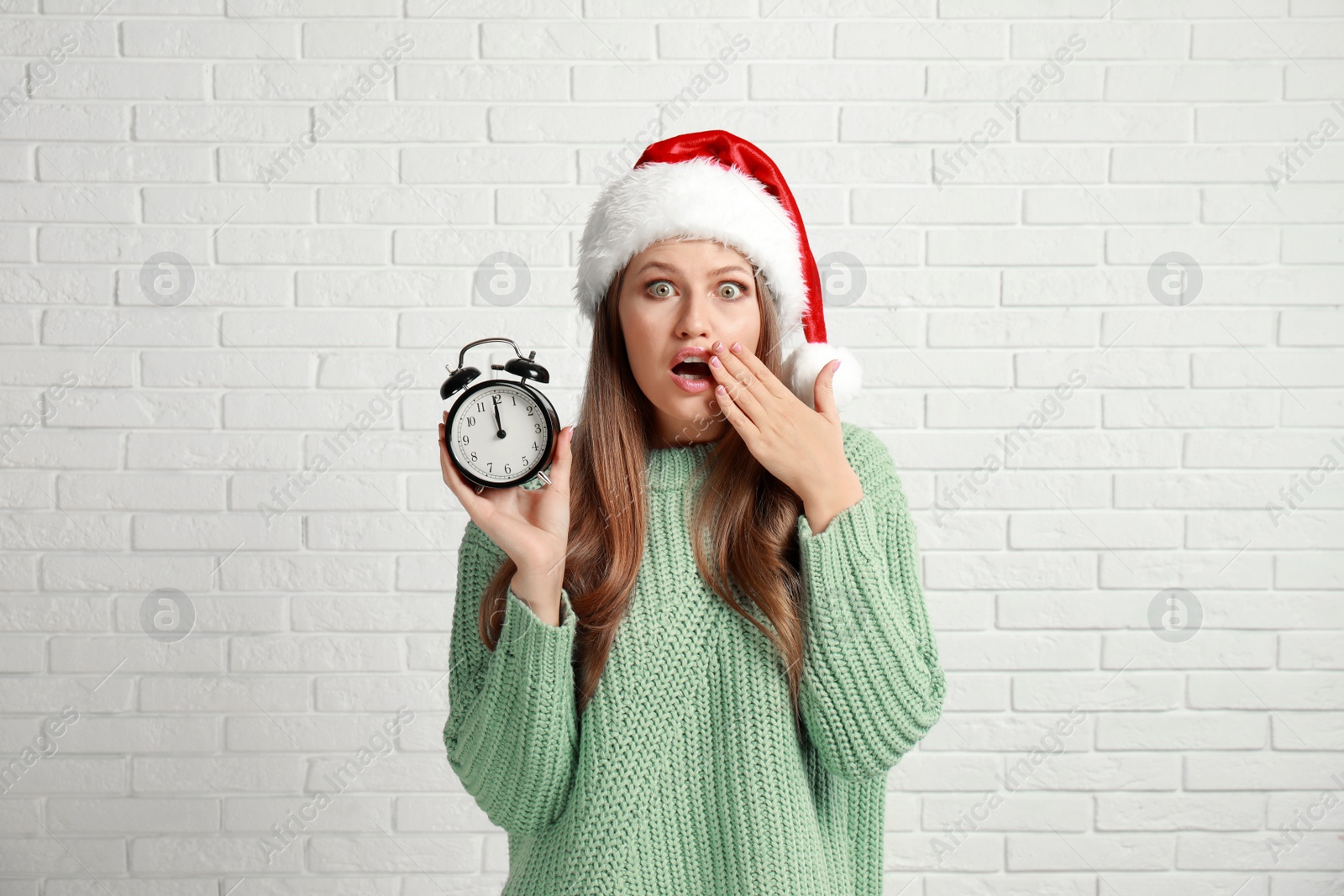 Photo of Emotional young woman in Santa hat with alarm clock near white brick wall. Christmas time