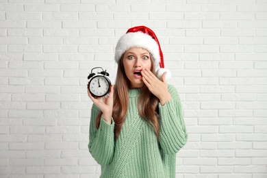 Photo of Emotional young woman in Santa hat with alarm clock near white brick wall. Christmas time