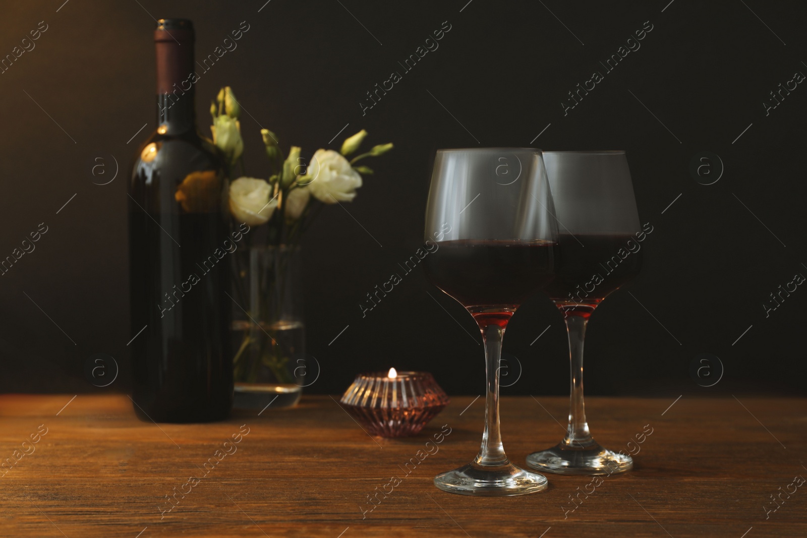 Photo of Glasses of wine, candles and flowers on wooden table near dark wall