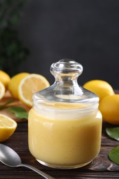 Photo of Delicious lemon curd in glass jar, fresh citrus fruits and spoon on wooden table