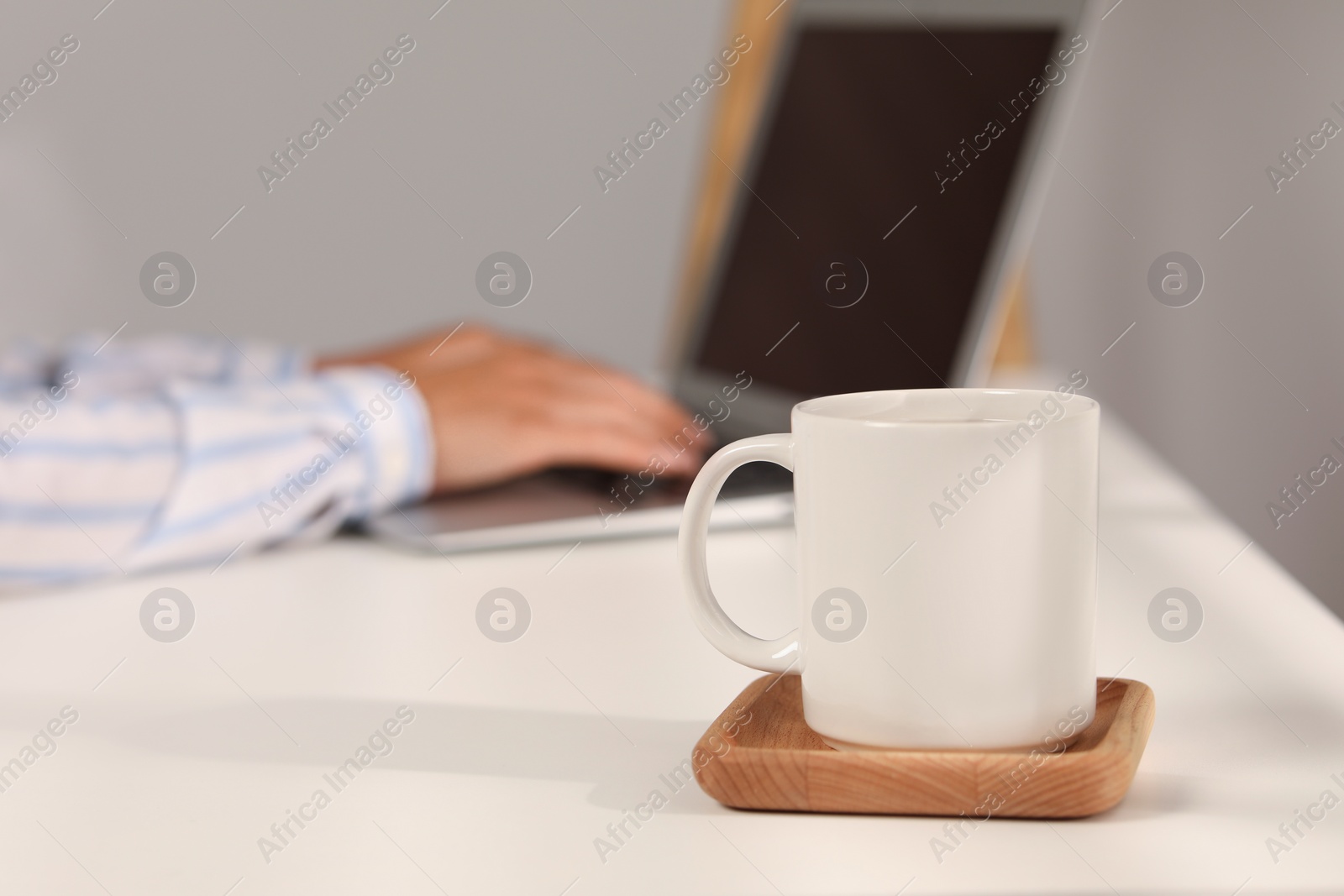 Photo of White ceramic mug on table, selective focus and space for text. Woman working with laptop at workplace, closeup