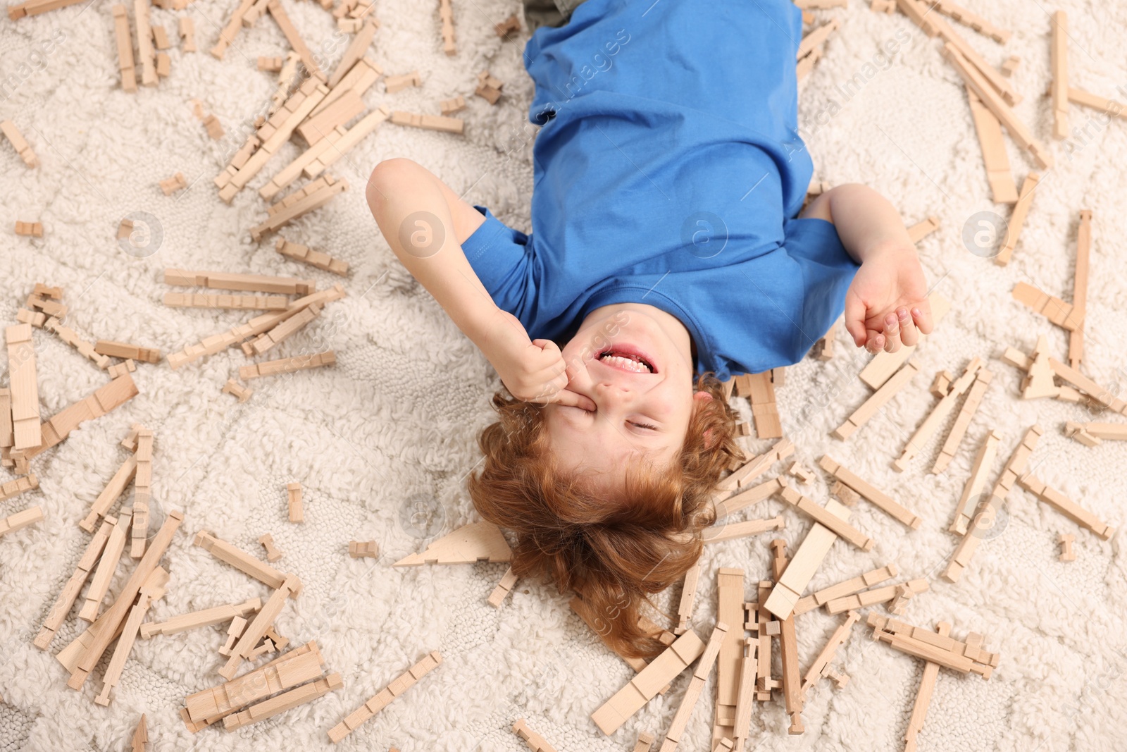 Photo of Cute little boy surrounded by wooden construction set on carpet at home, top view. Child`s toy