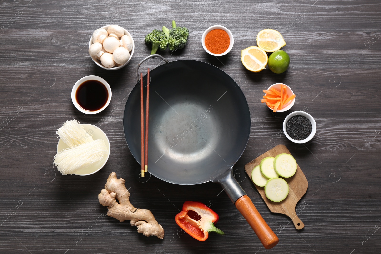 Photo of Empty iron wok and chopsticks surrounded by ingredients on dark grey wooden table, flat lay