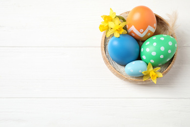 Photo of Colorful Easter eggs and flowers in bowl on white wooden table, top view. Space for text