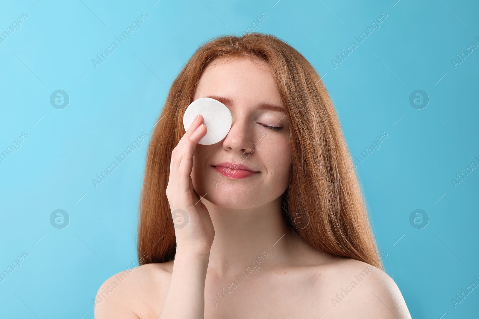 Photo of Beautiful woman with freckles covering eye with cotton pad on light blue background