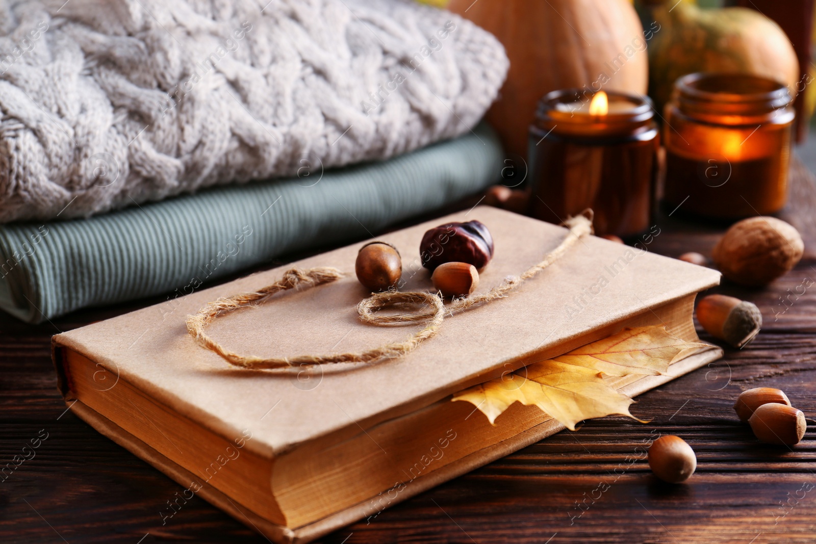 Photo of Book with autumn leaf as bookmark, scented candles and warm sweaters on wooden table