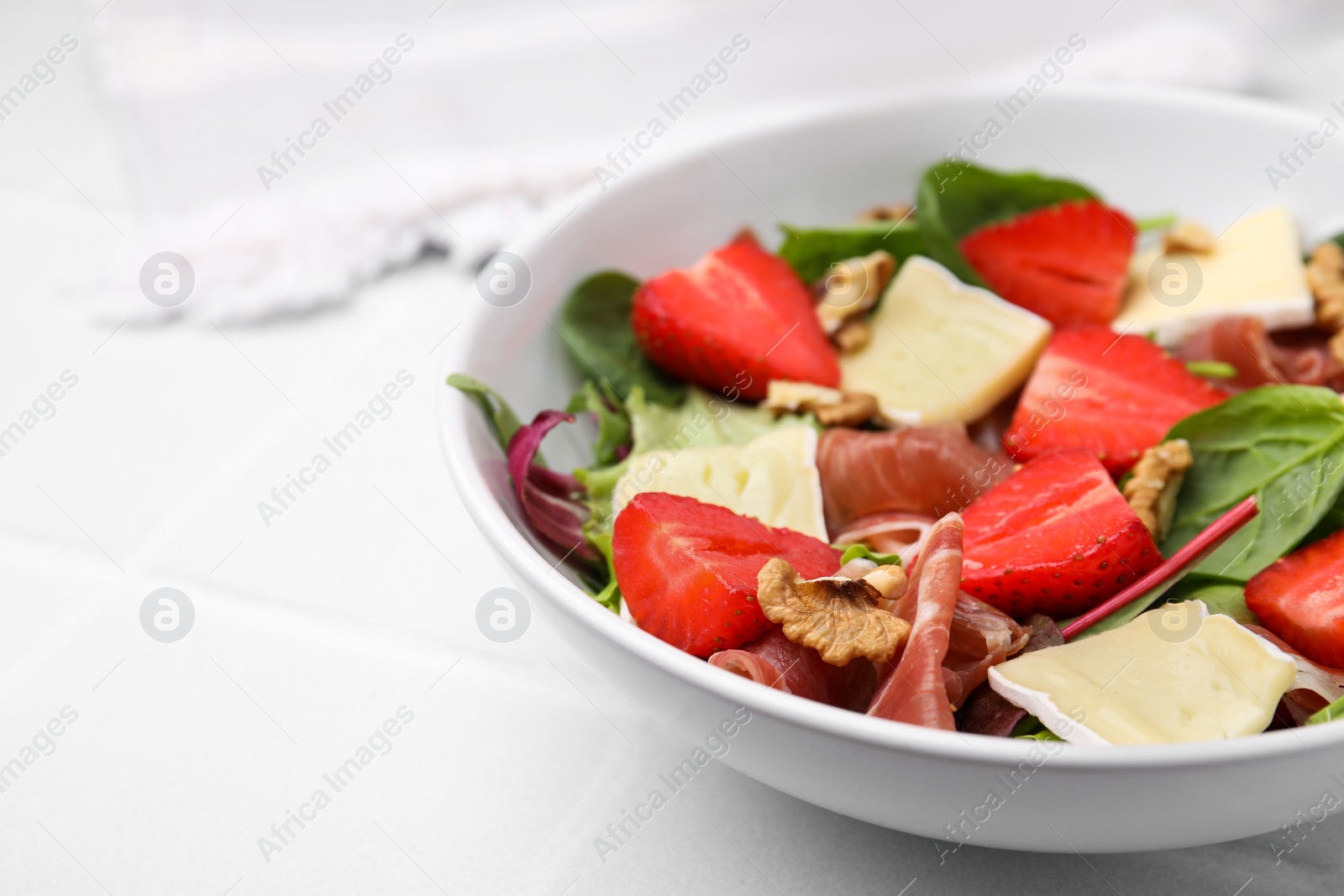 Photo of Tasty salad with brie cheese, prosciutto, strawberries and walnuts on white table, closeup. Space for text