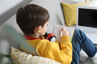 Photo of Cute little boy with headphones and modern laptop studying online at home. E-learning