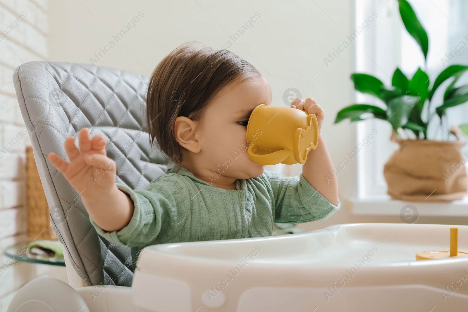 Photo of Cute little baby drinking from cup in high chair indoors