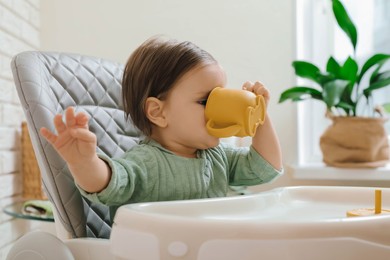 Cute little baby drinking from cup in high chair indoors