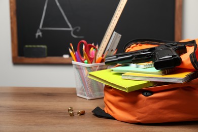 Photo of Gun, bullets and school stationery on wooden table near blackboard indoors. Space for text